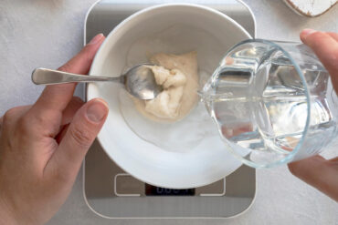 Eine weiße Schüssel auf einer Küchenwaage, ein Löffel hält eine Portion helles Anstellgut, aus einem Glas wird Wasser dazugegossen