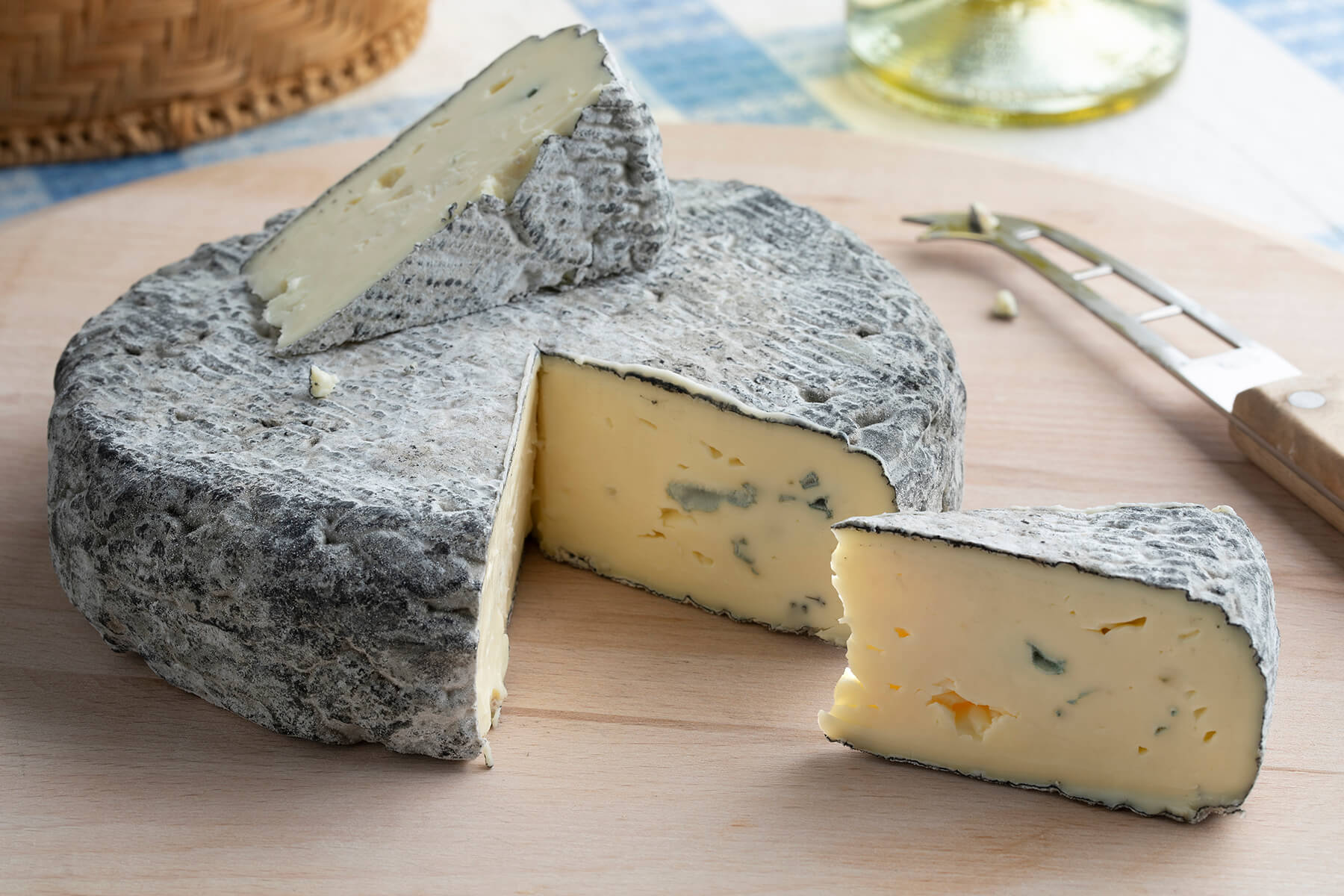 Runder Blauschimmelkäse mit grauer Ascherinde, ein Keil ist herausgeschnitten, ein Stück liegt obenauf und eines davor, im hellen, cremigen Teig wenige blaue Adern, daneben ein Käsemesser