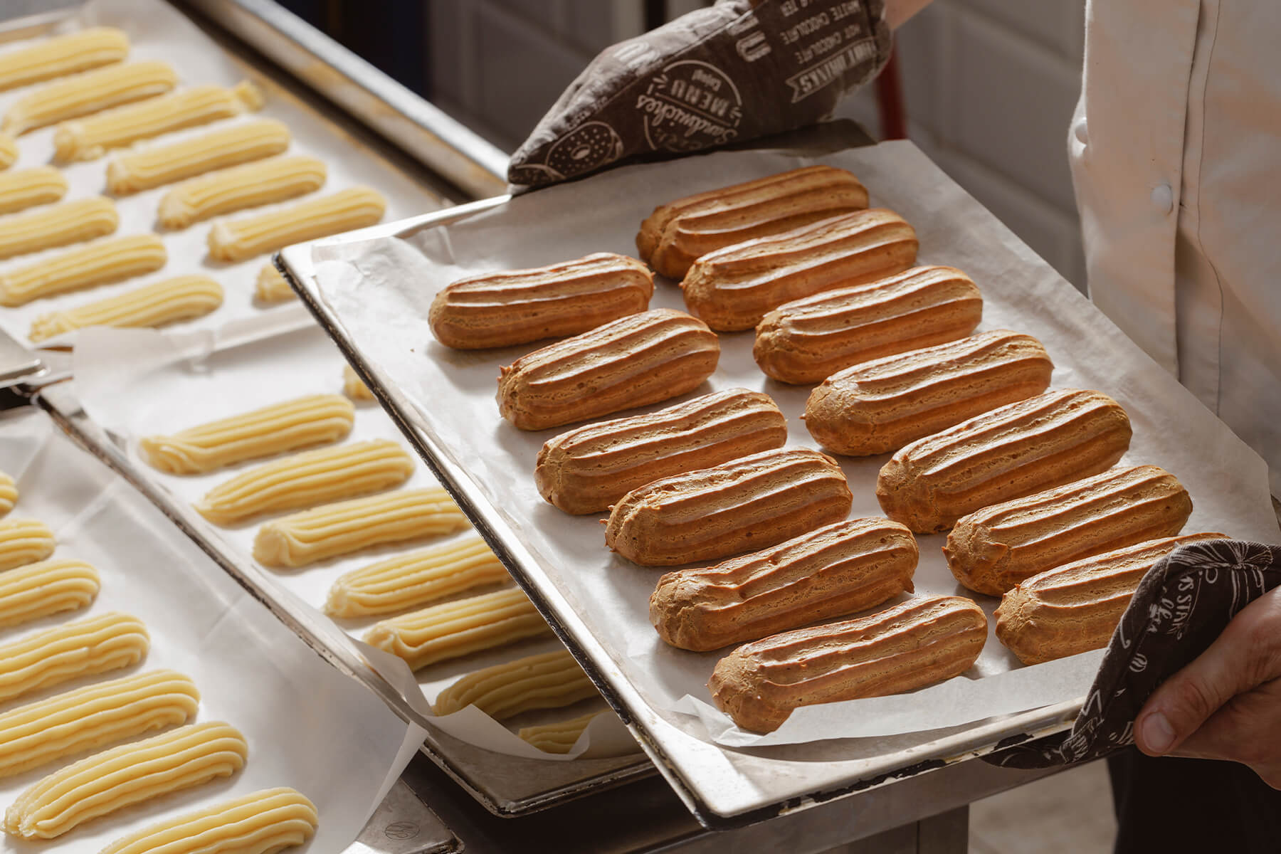Hände in gemusterten Ofenhandschuhen halten ein Blech mit goldbraun gebackenen Éclairs, dahinter Bleche mit rohen Teigstreifen