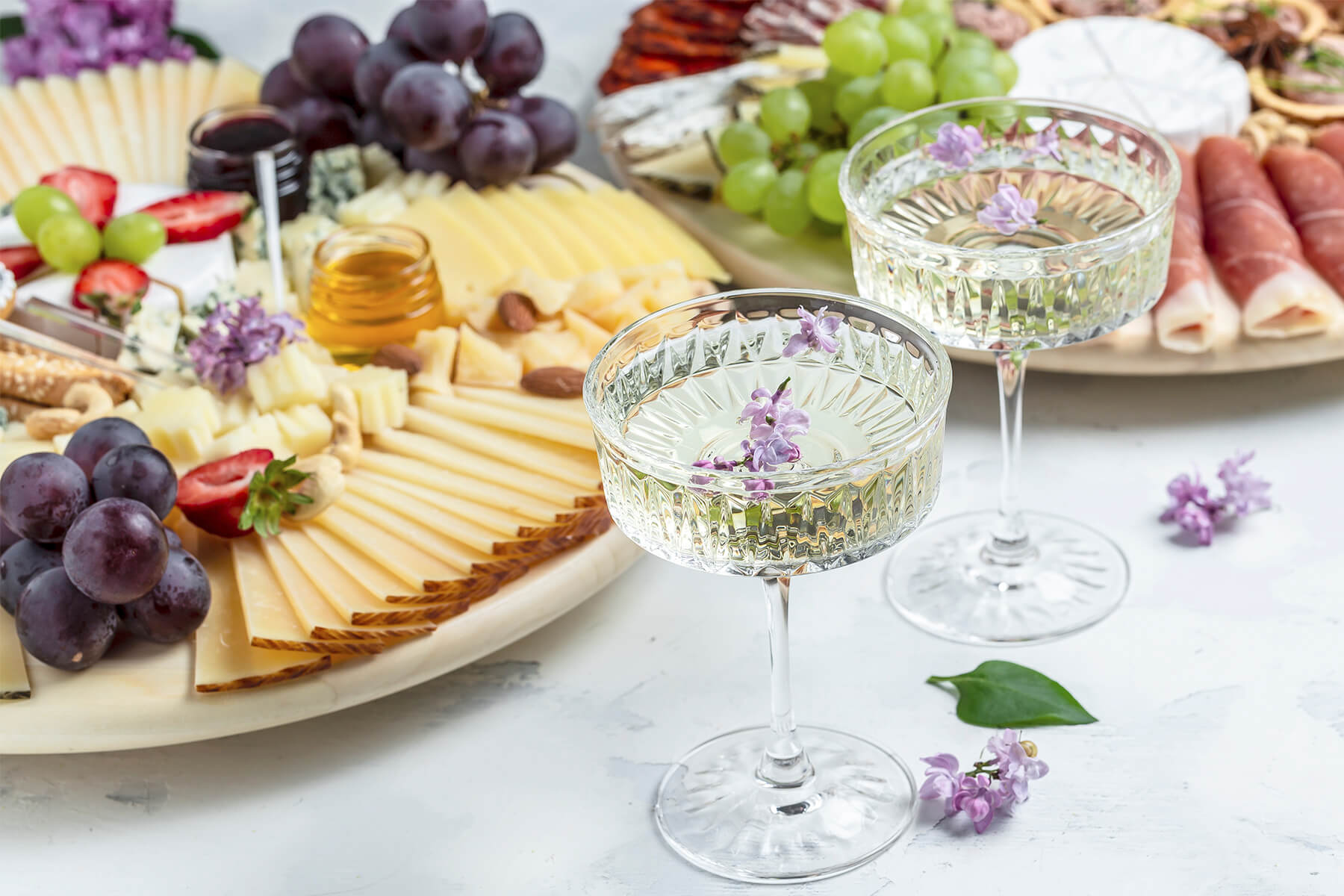Runde helle Platte schräg von vorn mit einem Fächer aus dünnen Schafskäsescheiben mit dunkler Rinde, Käsewürfeln, Gläschen mit Honig und Konfitüre, Trauben und Erdbeeren, im Vordergrund zwei Sektschalen mit Fliederblüten, dahinter eine zweite Platte mit Schinkenröllchen