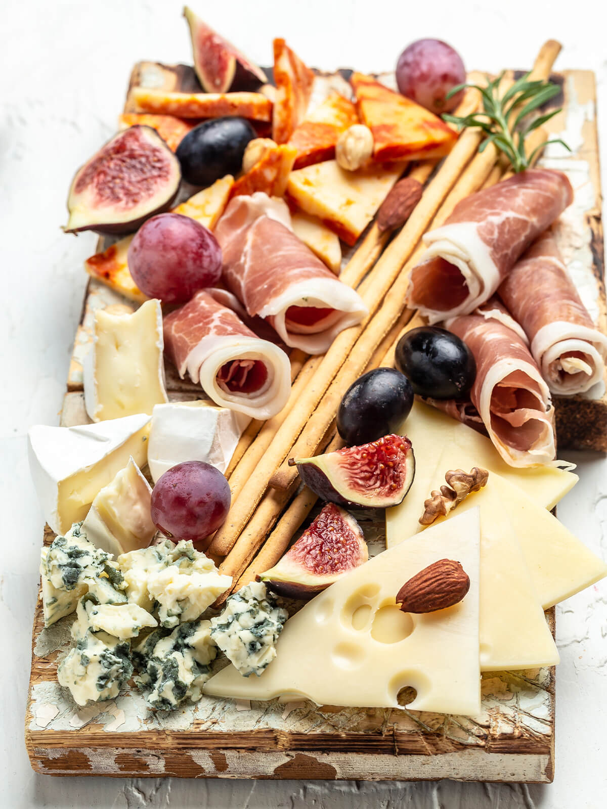 Rechteckiges Holzbrett von oben: Blauschimmelkäse, Brie-Spalten, Käsescheiben mit Löchern und ein Gewürzkäse in eigenen Ecken, dazwischen Schinkenröllchen, Grissini, Feigen und Trauben
