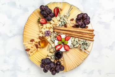 Runde Holzplatte von oben mit sechs Käsesorten: Camembert in Segmenten in der Mitte, Fächer aus dünnen Käsescheiben, Blauschimmelkäse auf Spießchen, Parmesanstücke, dazu Trauben, Erdbeeren, Honig und Sesamstangen