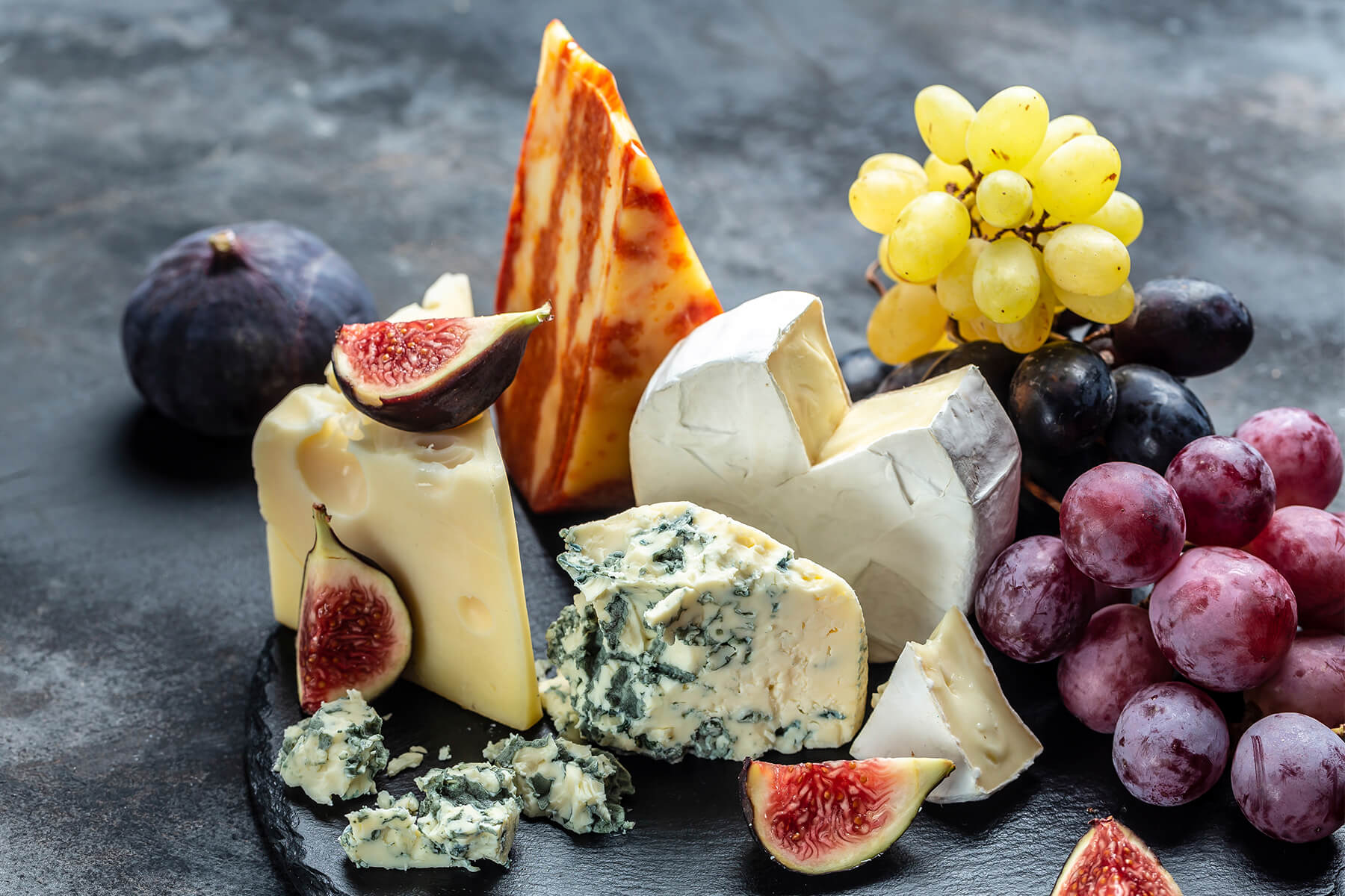 Vier Käsesorten auf einer runden Schieferplatte: Käse mit Löchern, rot marmorierter Käse, Brie mit weißer Rinde und Blauschimmelkäse, dazu Feigen und dreierlei Trauben