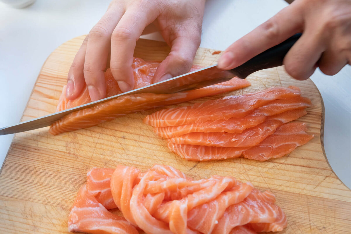 Roher Lachs wird auf einem Holzbrett mit einem langen Messer in dünne Scheiben geschnitten