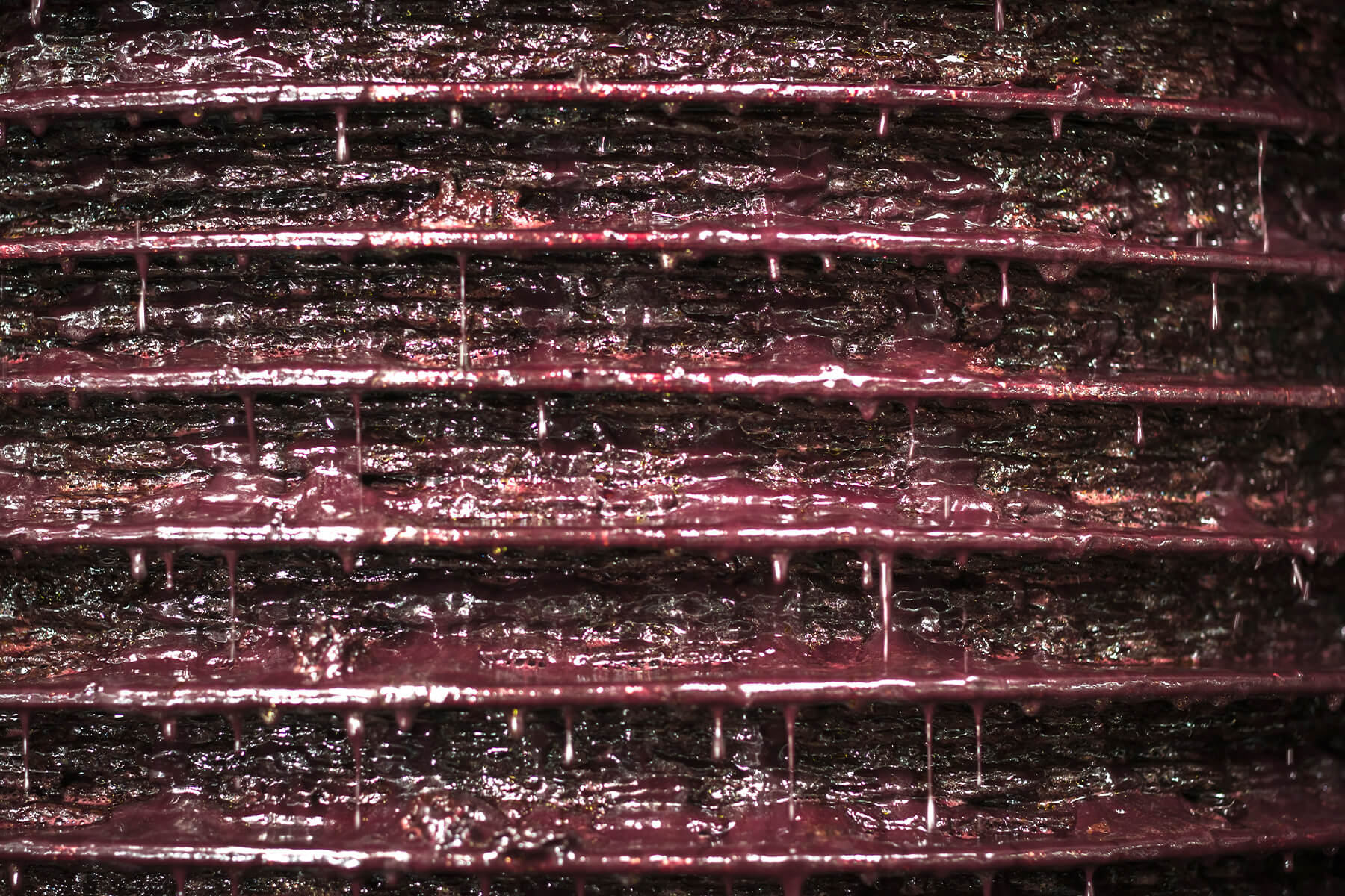 Nahaufnahme eines Stapels Pressmatten mit dunkler Olivenpaste, an jeder Mattenkante läuft rötlich schimmernde Flüssigkeit heraus und tropft nach unten