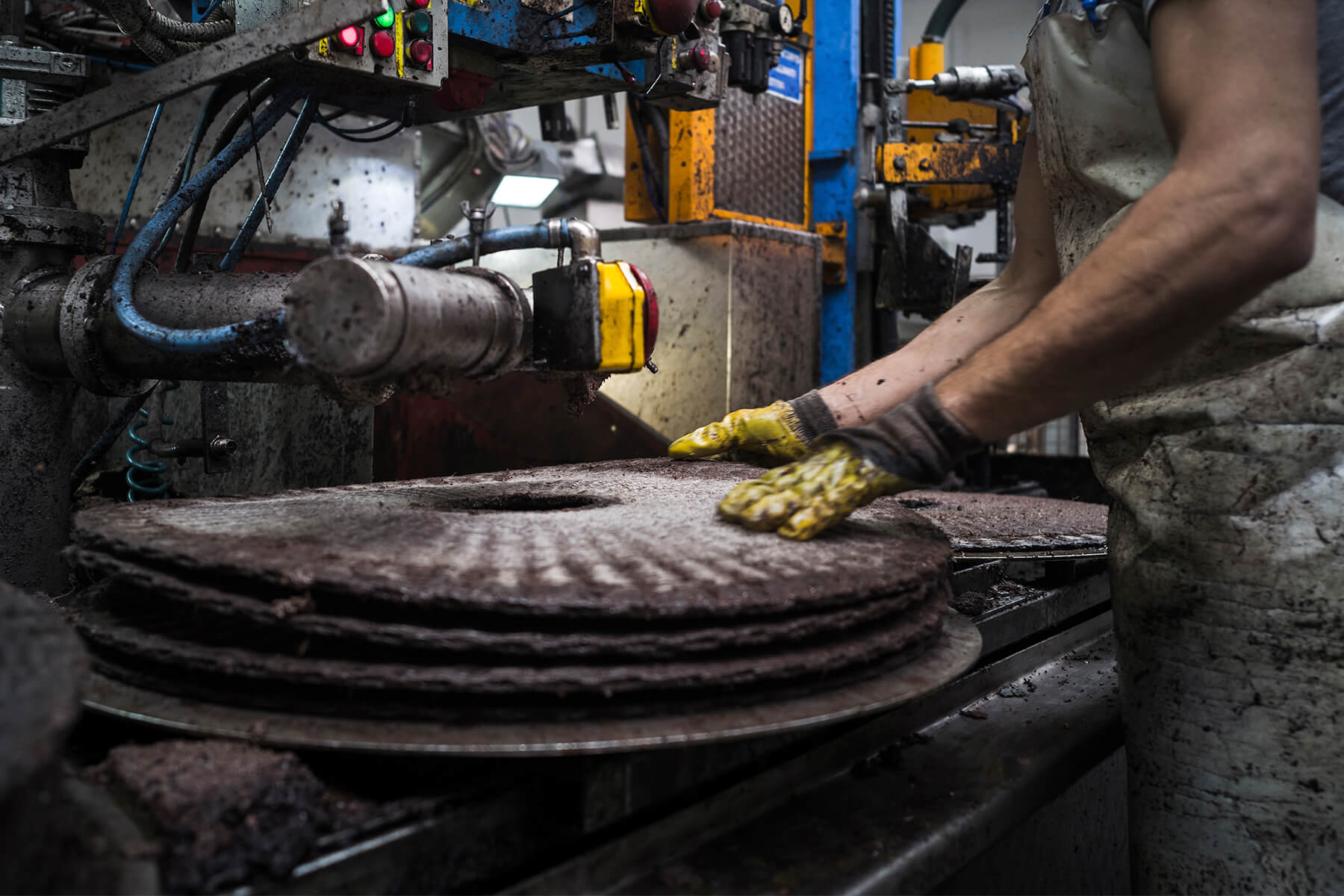 Ein Arbeiter mit Schürze und gelben Handschuhen legt runde Pressmatten mit dunkler Olivenpaste an der hydraulischen Presse übereinander