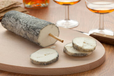 Ziegenkäserolle Sainte-Maure de Touraine mit grauer Ascherinde und einem Strohhalm in der Mitte auf einem Holzbrett, davor drei Scheiben mit weißem Teig, im Hintergrund zwei Gläser
