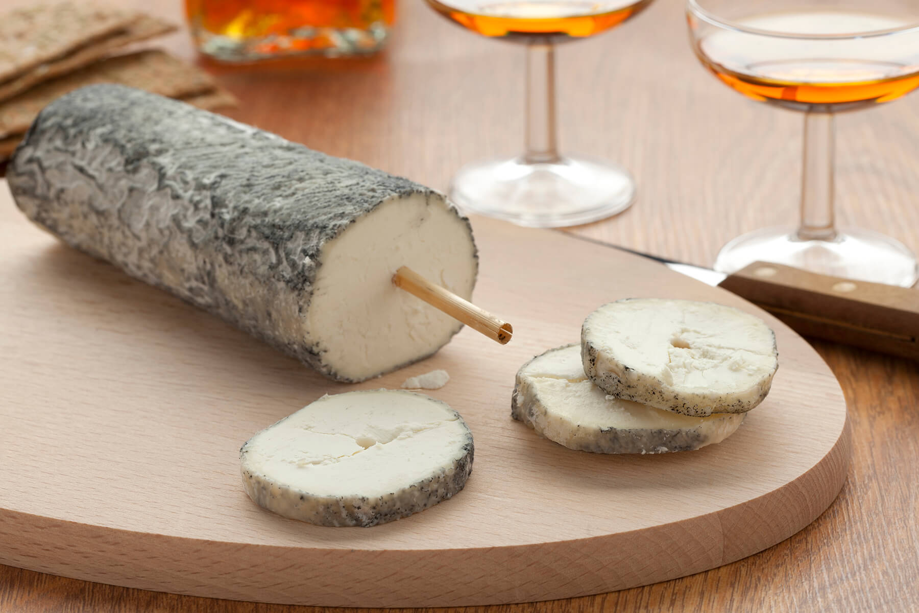 Ziegenkäserolle Sainte-Maure de Touraine mit grauer Ascherinde und einem Strohhalm in der Mitte auf einem Holzbrett, davor drei Scheiben mit weißem Teig, im Hintergrund zwei Gläser