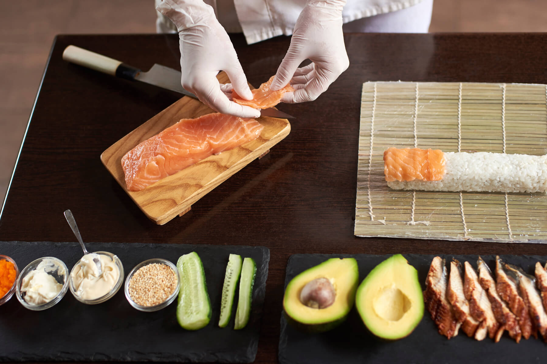 Hände in Handschuhen schneiden Lachs, daneben eine Bambusmatte mit Reis und Lachsstreifen sowie Gurke, Avocado und Sesam